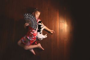 Girl in a red kimono-style dress lies on a dark wooden floor holding a black lace fan beside a sleeping black and white puppy.