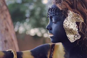 Profile of a person with dark blue and gold body paint, a beaded headpiece and gold ornament, with soft blurred trees behind.
