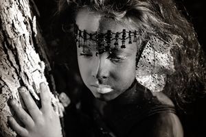 A black and white portrait of a child with a beaded headpiece and painted face leaning thoughtfully against a tree trunk.