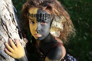Child with black and gold face paint, a beaded lace headpiece and curly hair leans against a tree trunk, looking up at the camera.