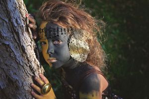 Woman with gold and black face paint, beaded headpiece and metallic ear ornament leans against a tree trunk in a forest.