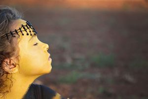 A child in profile with a gold-painted face and beaded headband tilts their head back in warm sunset light over an open field.