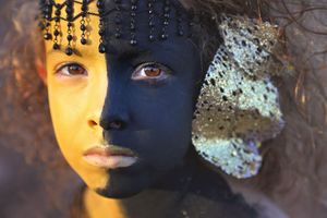 Close-up portrait of a child with split gold and black face paint, black beaded headdress and lace ornament in a creative photo.