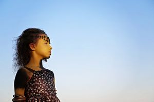 Young girl in a floral top and beaded headband gazes into the distance against a clear blue sky at golden hour.