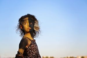 Child with curly hair and artistic face paint stands against a clear blue sky in warm golden light.