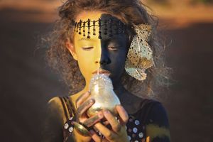 Young girl with half gold and half black face paint and beaded headpiece gazes down at a glowing glass jar in warm light.