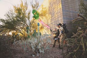 Two people in Ghostbusters-style costumes with proton packs aim glowing green and pink streams near a building in a desert garden.