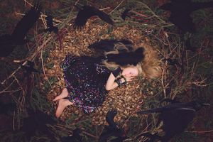 Girl in a sequined dress sleeps barefoot on a nest of dry leaves while black crow shadows surround her in the forest.