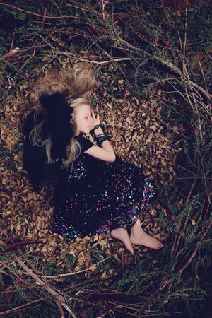 Blonde girl with black feathered wings lies asleep on brown leaves in a sparkly dress, viewed from above in a wooded setting.