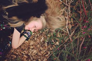 Close-up of a sleeping blonde girl with black wings and beaded bracelets resting on autumn leaves among wild twigs.