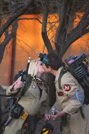 A couple in tan Ghostbusters jumpsuits with proton packs share a kiss in front of glowing orange light and bare trees.