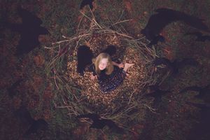 Overhead view of a girl lying in a circular leaf nest with arms outstretched as dark crow shapes surround her.