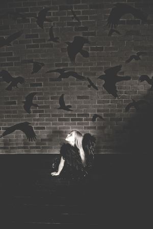 Black and white photo of a girl with dark wings sitting on the floor, gazing up at crow silhouettes on a brick wall.