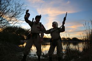 Two people in Ghostbusters-style jumpsuits and proton packs raise their arms beside a lake at sunset.