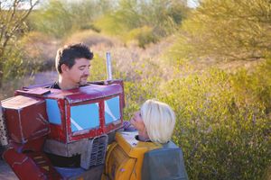 Man in a handmade red and blue robot costume smiles beside a blonde woman in a yellow robot costume in a sunny outdoor field.