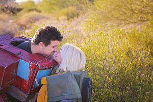 A smiling man leans over a red toy-style vehicle to kiss a blonde woman among golden desert brush at sunset.