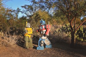 Two handmade Transformers costumes, one green and yellow and one red and blue, stand facing each other beside desert cacti.