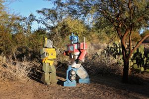 Two robot sculptures resembling Transformers stand among desert trees and cactus under a bright blue sky.