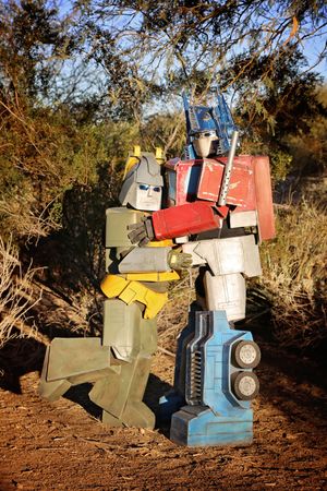 Two cardboard-style Transformers costumes in red, blue, green and yellow embrace on a dirt trail surrounded by desert trees.