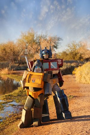 Costumed Optimus Prime and Bumblebee-style Transformers pose on a dirt path beside a small pond under a blue sky.