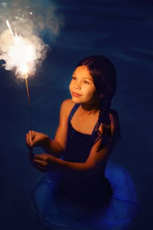 A smiling girl in a blue dress stands in dark water holding a sparkler that releases glowing smoke and light.
