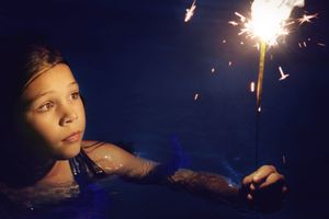 A young girl in a dark pool gazes at a bright burning sparkler, with glowing sparks lighting her face at night.