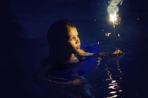 A girl swims in dark nighttime water while holding a sparkler, its fiery glow reflecting on the rippling surface.