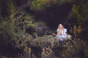 A woman dressed as Alice in Wonderland sits quietly in a dark, overgrown forest surrounded by wild shrubs and yellow flowers.