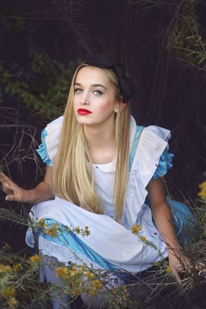 A blonde woman with red lipstick, dressed as Alice in Wonderland in a blue and white apron dress, sits among yellow wildflowers.