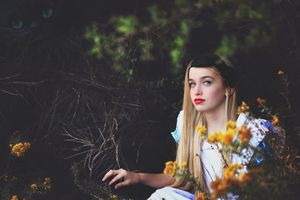 Young woman in a light blue dress with red lipstick sits among yellow wildflowers in a dark forest, with a faint Cheshire Cat face above.