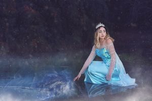 A woman in a sparkling blue gown and tiara kneels beside misty reflective water in a magical, wintry fairy-tale scene.