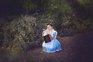 Girl dressed as Alice in a blue dress and white apron kneels on a woodland path, peering into a black top hat among golden shrubs.