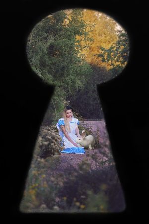View through a keyhole of an Alice in Wonderland cosplayer in a blue dress kneeling beside a white rabbit on an autumn garden path.