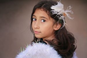 Young girl with long dark wavy hair and a white feather hair clip looks over her shoulder, wearing a fluffy white feather boa.