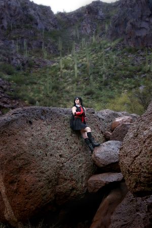 Cosplayer in a black skirt, red top and boots sits on a large boulder in a desert canyon dotted with saguaro cacti.