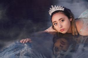 Young woman wearing a sparkling silver tiara rests her arms on an icy reflective surface, her reflection mirrored in the dark water below.