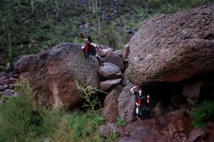Two cosplayers in black and red outfits climb among large desert boulders surrounded by green brush and cacti.