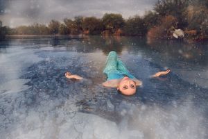 Woman in a turquoise dress floats on her back on a calm, icy lake surrounded by autumn trees under a cloudy, moody sky.