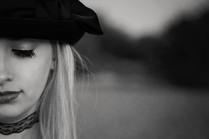 Black and white close-up of a blonde woman in a black hat and lace choker, her eyes closed, with a softly blurred landscape behind her.