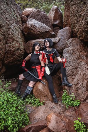 Two cosplayers in black dresses with red gloves and scarves pose with swords on large red rocks surrounded by green plants.