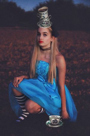 Alice in Wonderland inspired portrait of a woman in a blue dress and striped socks balancing a stack of teacups on her head.