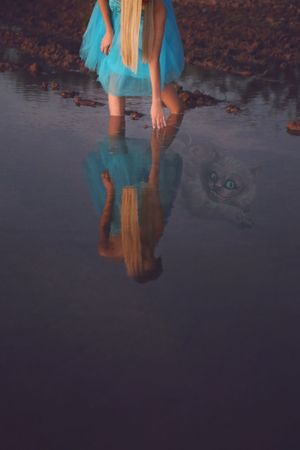 Girl in a blue tulle dress with long blonde hair bends to touch still water, her reflection and a ghostly Cheshire Cat visible below.