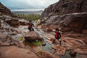 Two cosplayers in black and red costumes explore rocky red terrain with shallow pools in a rugged canyon landscape.