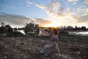 Woman in a flowing vintage dress reaching toward the glowing sunset over a misty lake, with dramatic clouds and trees.