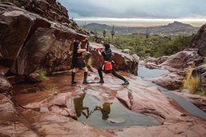 Two cosplayers in black and red costumes face off with swords on wet red rock slabs with a mountain and cacti behind.