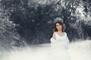 Young woman in a white gown and sparkling tiara standing in a snowy, frosted forest as snowflakes fall around her.