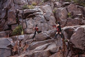 Two cosplayers in dark outfits with red accents stage a sword fight on a steep rocky cliff face.