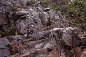 Two cosplayers in black and red costumes climb across a rugged red rock cliff in a desert landscape.