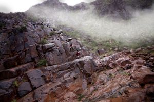 A tiny cosplayer in red and black stands on a vast rocky slope beneath drifting mist and low clouds.