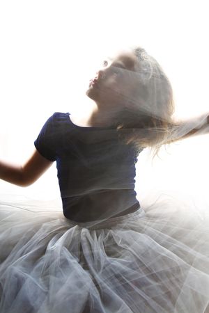 Young girl in a navy top and gray tulle skirt twirls in bright backlight, with soft motion blur and flowing hair.